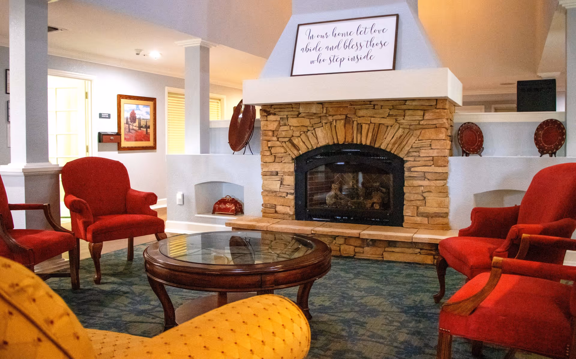 A cozy living room area with a stone fireplace in the center. The fireplace has a sign on top that reads, 'In our home let love abide and bless those who step inside.' Surrounding the fireplace are red upholstered armchairs and a round wooden coffee table with a glass top. The room has a blue patterned carpet and light-colored walls with some framed artwork and decorative plates.