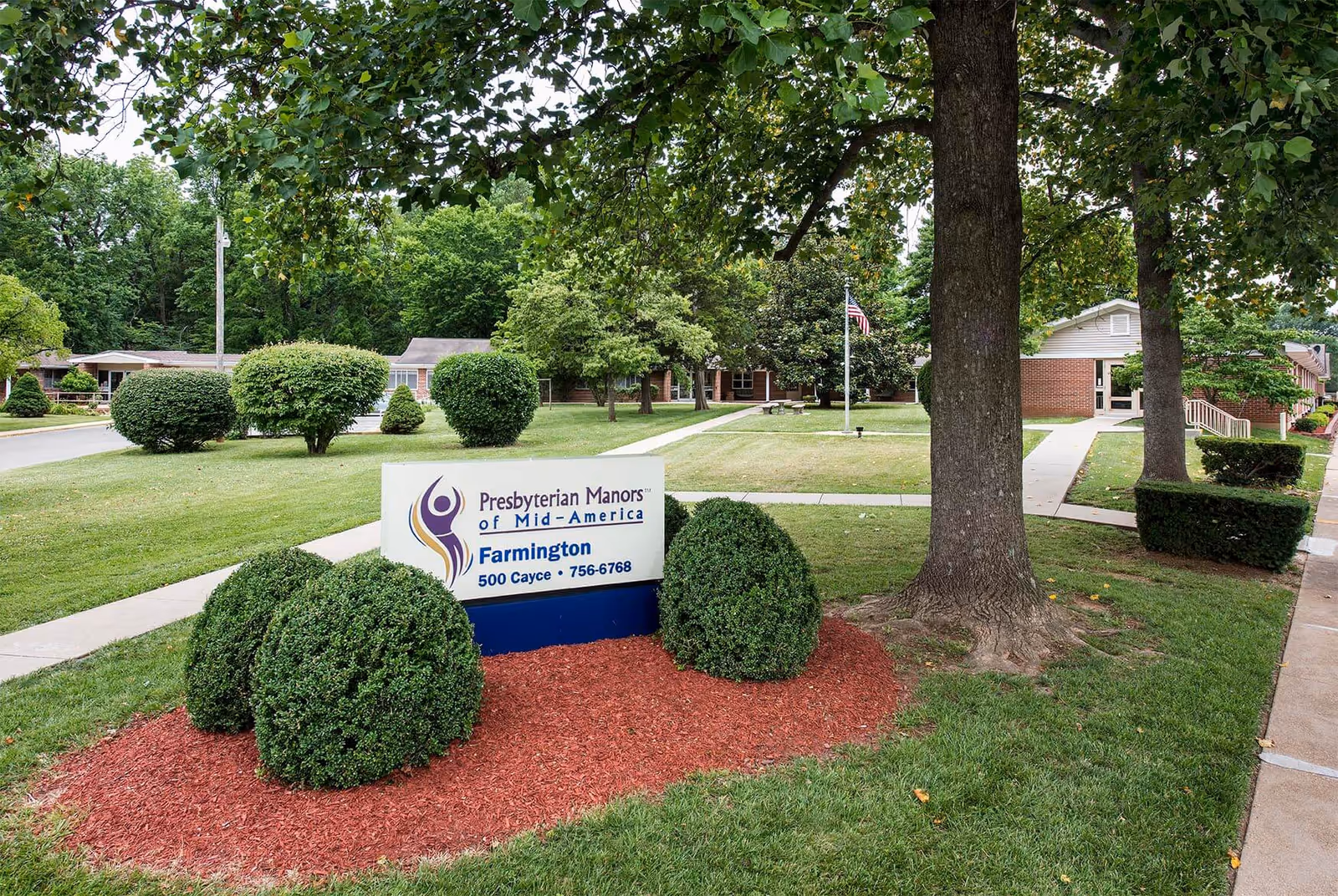 Front lawn and walkways of Farmington Presbyterian Manor with a sign, trimmed bushes, large trees, and an American flag near the building.