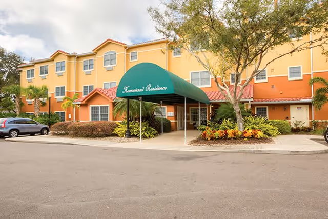 Front entrance of a three-story yellow and orange senior living building with a green canopy, landscaping, and parked cars.