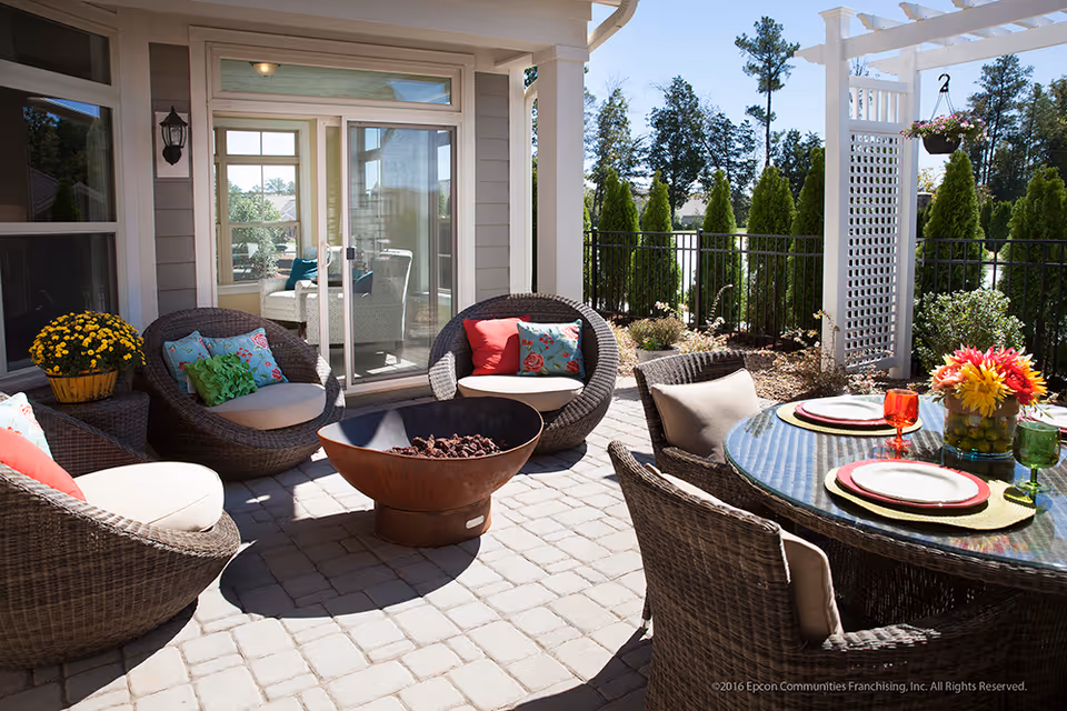 Outdoor patio area with wicker chairs and a round glass table set with plates and colorful glasses. There is a fire pit in the center and potted plants around the seating area. The patio is adjacent to a building with sliding glass doors and windows, surrounded by greenery and trees.