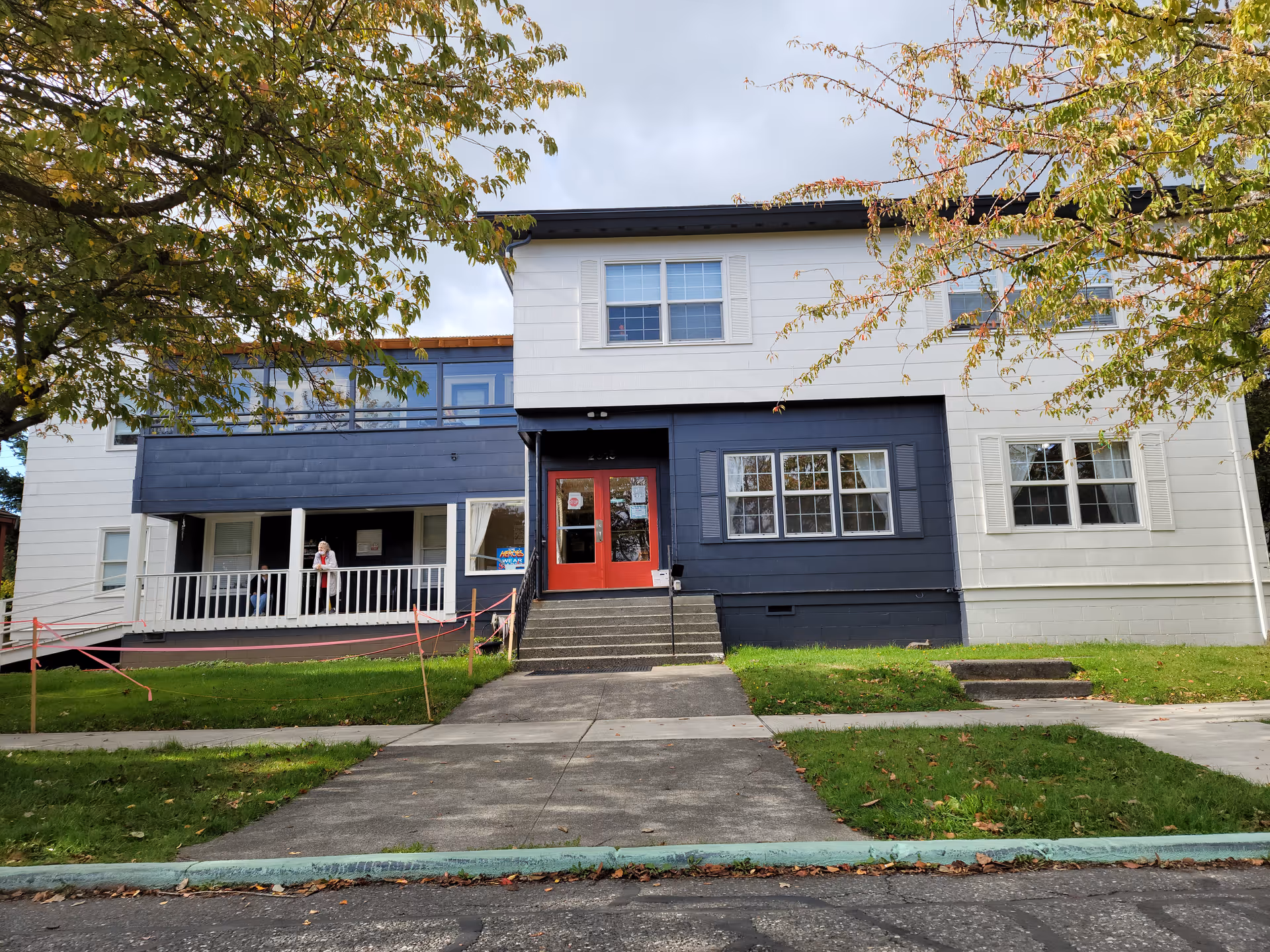 Front exterior view of a two-story building with white and dark blue siding, red double doors at the entrance, and a small porch with railings. There are trees with green and some autumn-colored leaves on either side of the building, and a concrete walkway leads up to the entrance.