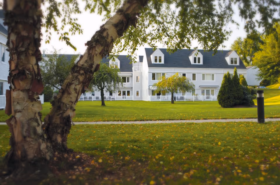 White multi-story senior living building with dormer windows seen across a grassy lawn framed by birch trees.