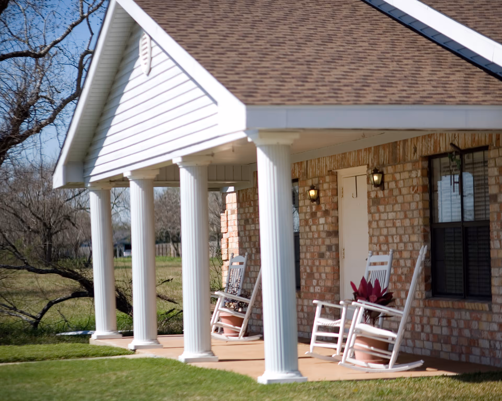 Front porch of a brick building with white columns and a brown shingled roof. Two white rocking chairs with potted plants are placed on the porch. The area around the building is grassy with leafless trees in the background.