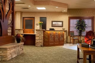 Reception area with a wooden front desk featuring carved leaf designs, stone accents, a vase with yellow flowers, and a framed picture on the wall behind. To the left is a stone bench with a potted plant, and to the right are chairs near a window with blinds and a tall potted plant. The room has carpeted flooring and a ceiling with recessed lighting.