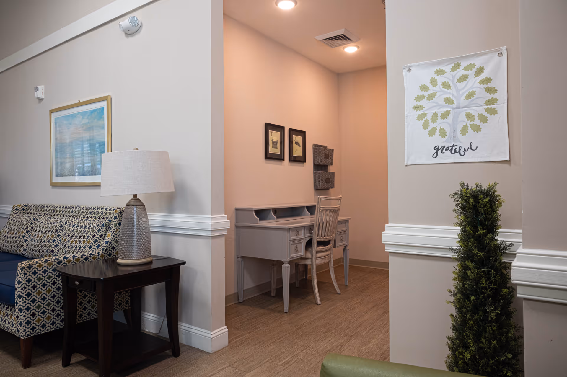 Interior view of a senior living facility showing a cozy seating area with a patterned couch and a side table with a lamp. In the background, there is a small desk with a chair placed in a recessed nook, decorated with framed pictures on the wall. A wall hanging with a tree and the word 'grateful' is visible, along with a tall green plant in the foreground.