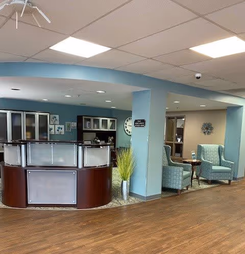 Interior view of a senior living facility reception area with a curved wooden front desk, light blue walls, and wood flooring. To the right, there are two light blue upholstered armchairs with a small wooden table between them. The ceiling has recessed lighting and a ceiling fan. A clock and some decorative items are visible on the walls.