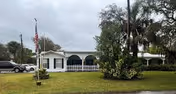 Single-story white building with a covered porch and arches, surrounded by green lawn and trees. Several parked cars are visible on the left side, and an American flag is flying on a flagpole in front of the building under a cloudy sky.