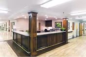 Reception desk area inside Pleasanton Nursing and Rehabilitation Center with wooden paneling and columns, a computer, and a framed picture on the wall behind the desk. The floor is light wood, and hallways extend on both sides of the reception area.