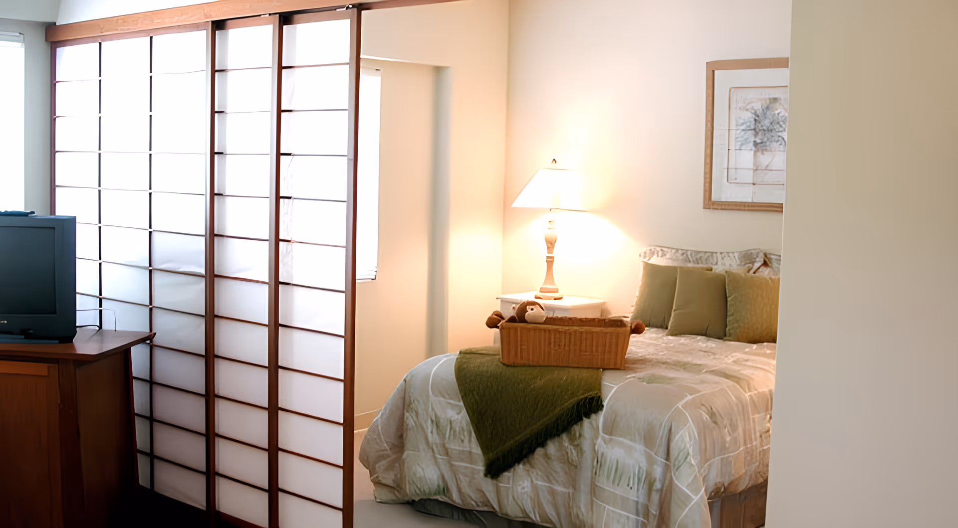 A cozy bedroom with a bed covered in a beige and green patterned comforter and green pillows. A wooden basket with a stuffed animal is placed on the bed. A lit table lamp sits on a nightstand next to the bed. A wooden TV stand with an old-style television is visible to the left, partially obscured by a white and wooden folding room divider.