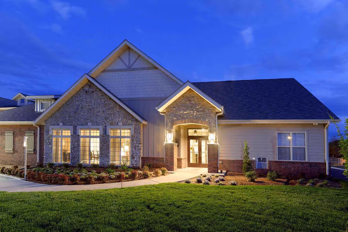 Exterior view of The Castlewood Senior Living building at dusk, featuring a well-lit entrance with stone and brick facade, large windows, and a neatly landscaped lawn and garden.