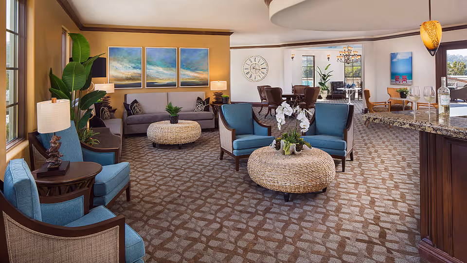 A spacious and well-lit living room area with blue upholstered chairs, a gray sofa, two round woven coffee tables, decorative plants, and framed artwork on the wall. The room features large windows, a patterned carpet, and a granite countertop bar area with wine glasses and a bottle of wine.