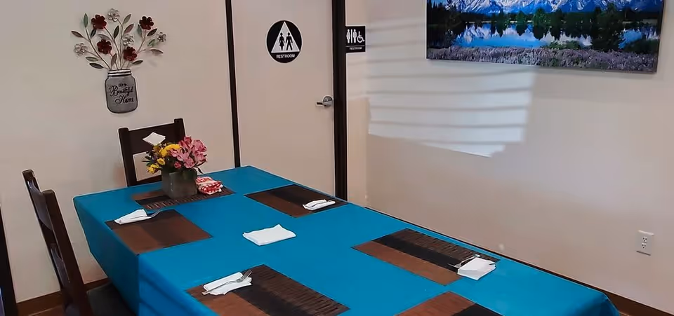 A dining table covered with a blue tablecloth set with four place mats and folded napkins. A small flower arrangement is in the center of the table. On the wall behind the table is a door marked as a restroom with accessibility signs, a decorative wall piece with flowers in a jar labeled 'Beautiful Home,' and a landscape painting.