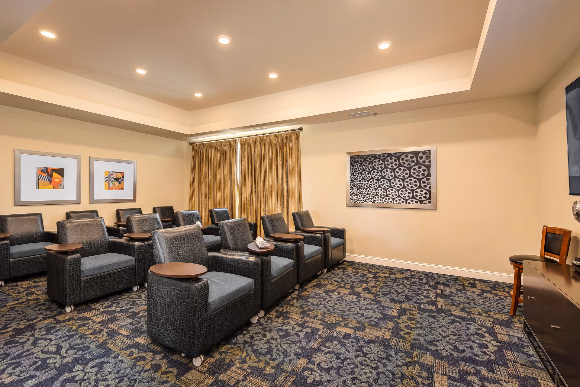 A small theater or media room with two rows of black cushioned chairs, each with a small attached wooden table. The room has patterned carpet, beige walls, framed artwork on the walls, and a window covered with gold curtains. There is a TV mounted on the right wall above a wooden cabinet with a chair next to it.