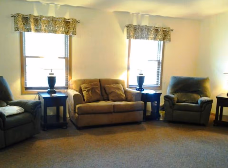 A cozy living room with two armchairs and a loveseat arranged around two side tables, each with a lamp. Two windows with patterned valances let in natural light, and the room has beige walls and carpeted flooring.