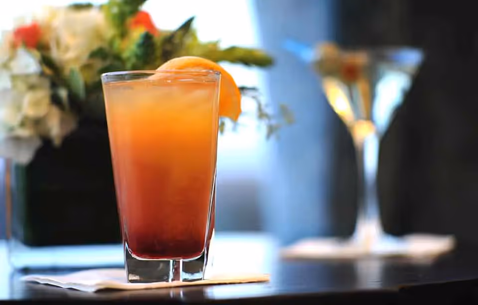 A close-up of a tall glass filled with a layered orange and red drink garnished with an orange slice, placed on a napkin on a table. In the blurred background, there are two martini glasses and a floral arrangement.