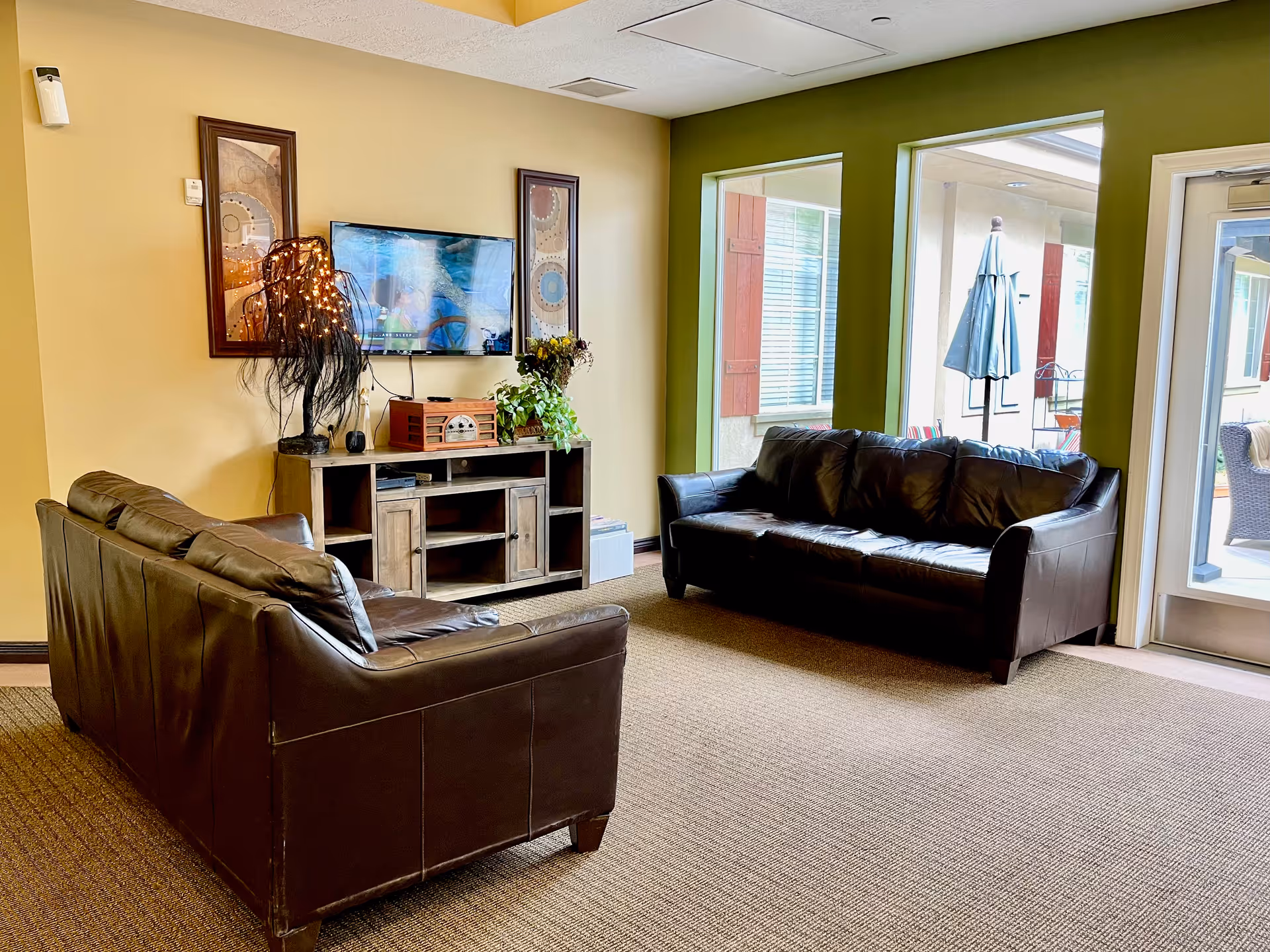 A cozy living room area with two dark brown leather sofas facing a wall-mounted flat screen TV. Below the TV is a wooden entertainment console with decorative items including a lamp with string lights, a vintage-style radio, and a potted plant. The walls are painted yellow and green, and there are two large windows and a glass door leading to an outdoor patio area with an umbrella and chairs.