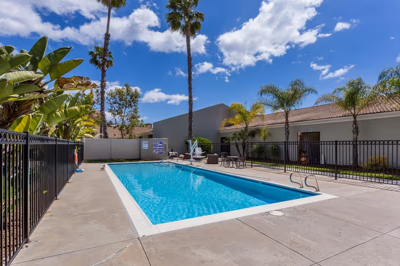 Outdoor swimming pool area at a senior living facility with clear blue water, surrounded by a concrete deck and black metal fencing. There are palm trees and other tropical plants around the pool, with a building in the background under a partly cloudy blue sky.