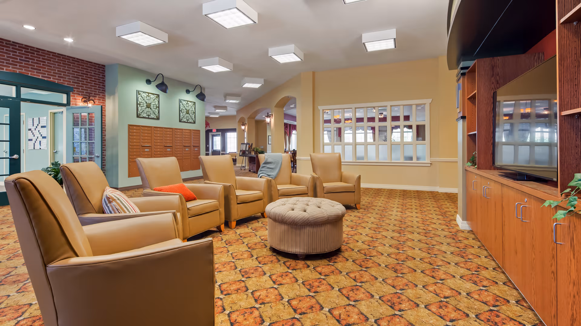 A spacious living room area in an assisted living facility with five tan leather armchairs arranged around a round tufted ottoman. The room has patterned carpet flooring, a large wooden entertainment center with a flat-screen TV, and decorative wall art. There are windows and doors in the background, along with soft lighting fixtures on the ceiling.