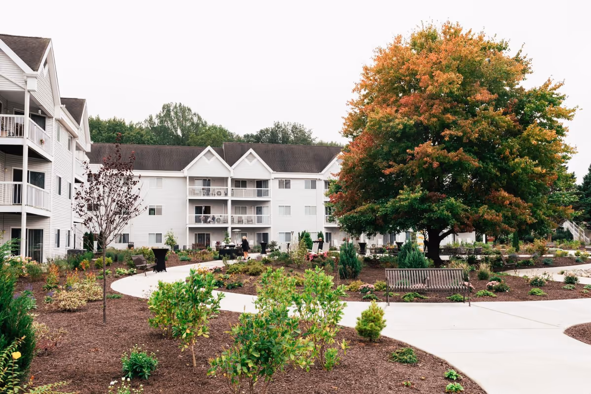 Outdoor garden area with a large tree showing early autumn colors, surrounded by landscaped plants and shrubs. There are benches and a winding concrete pathway. In the background, there is a white multi-story residential building with balconies.