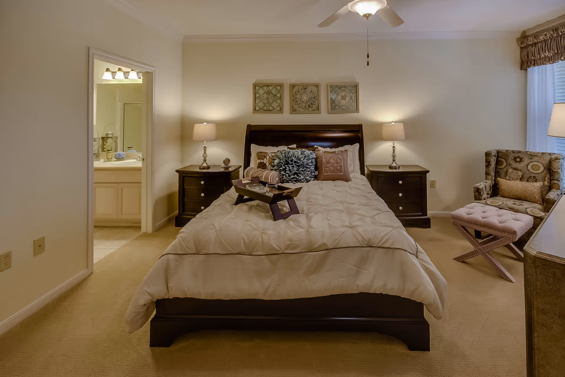 A cozy bedroom with a neatly made bed featuring multiple decorative pillows and a tray on top. There are two matching dark wood nightstands with lamps on either side of the bed. Three framed decorative artworks hang above the bed. To the right, there is a patterned armchair with a small cushioned ottoman in front of it, next to a window with curtains and a floor lamp. An open doorway on the left side of the room leads to a bathroom with a sink and mirror visible.