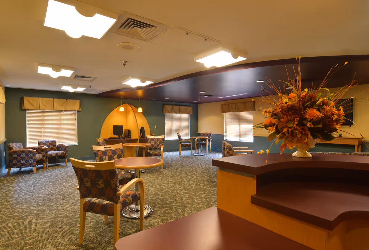 Spacious senior living common room with multiple patterned chairs and tables, a reception counter with a large floral arrangement, and a computer station in the back.