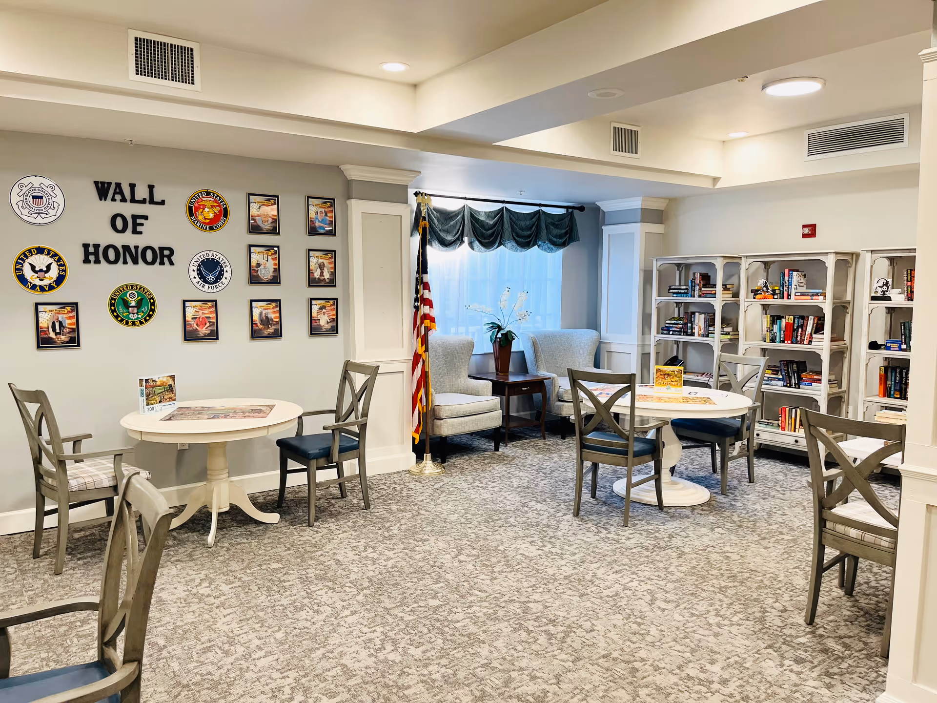 Bright communal lounge with round tables and chairs, bookshelves, an American flag, and a 'Wall of Honor' display on the wall.