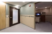 Interior corridor of a senior living facility with a frosted-glass door on the left and an alcove containing a TV and cabinet on the right.