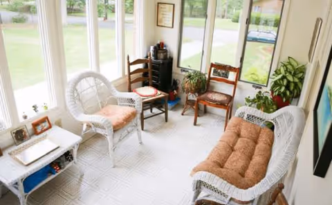 Sunlit enclosed seating area with wicker chairs, a tufted sofa, and potted plants.