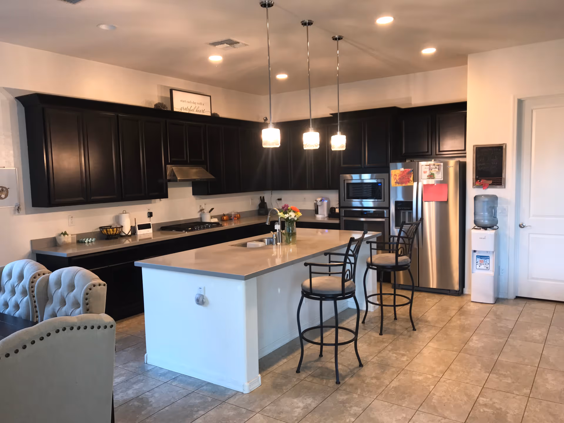 Bright modern kitchen with a large island topped by three pendant lights, bar stools, dark cabinets, and stainless steel appliances.