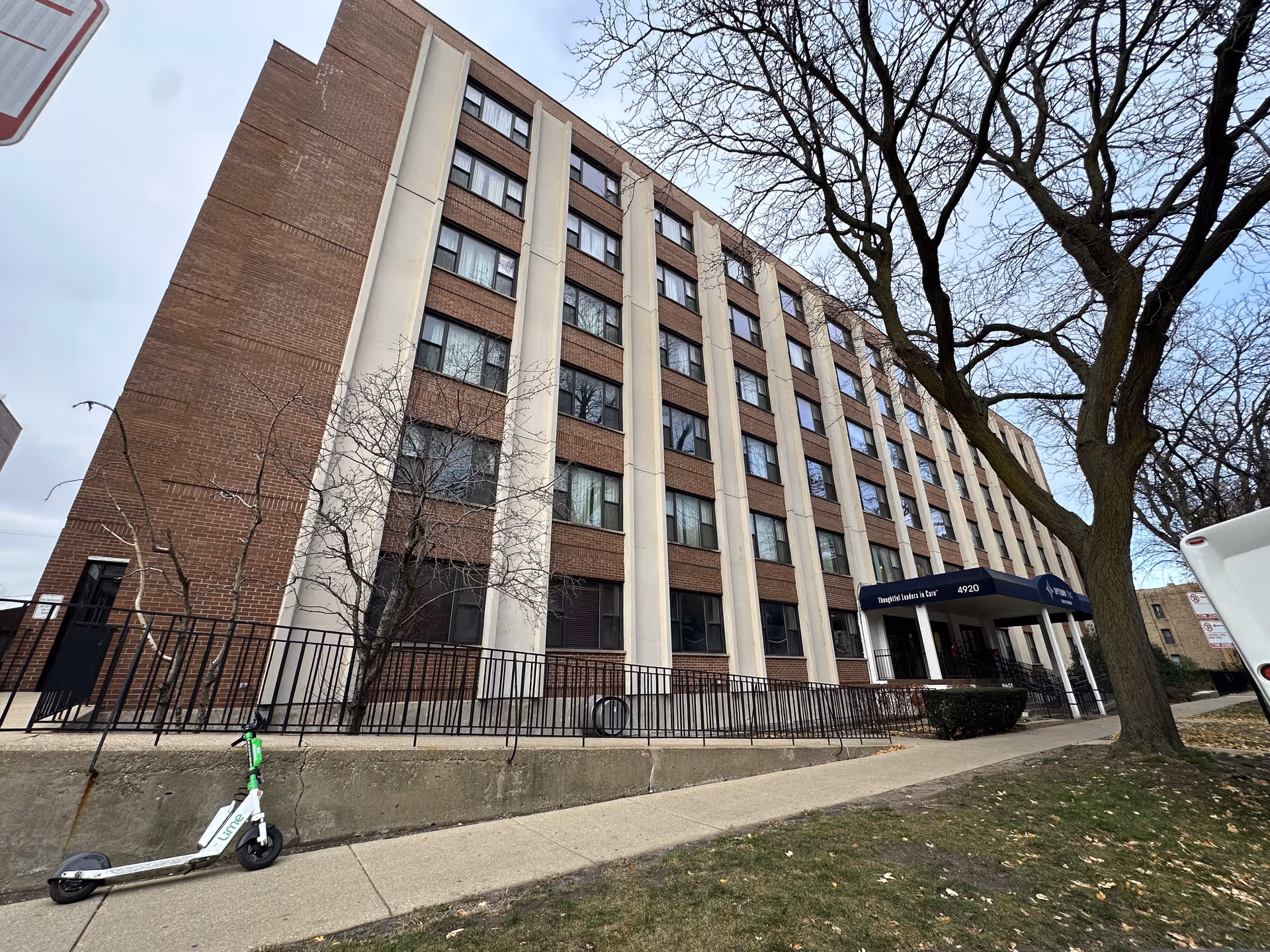 Exterior view of a multi-story brick-and-concrete care facility with an entrance canopy, leafless trees, and a scooter on the sidewalk.