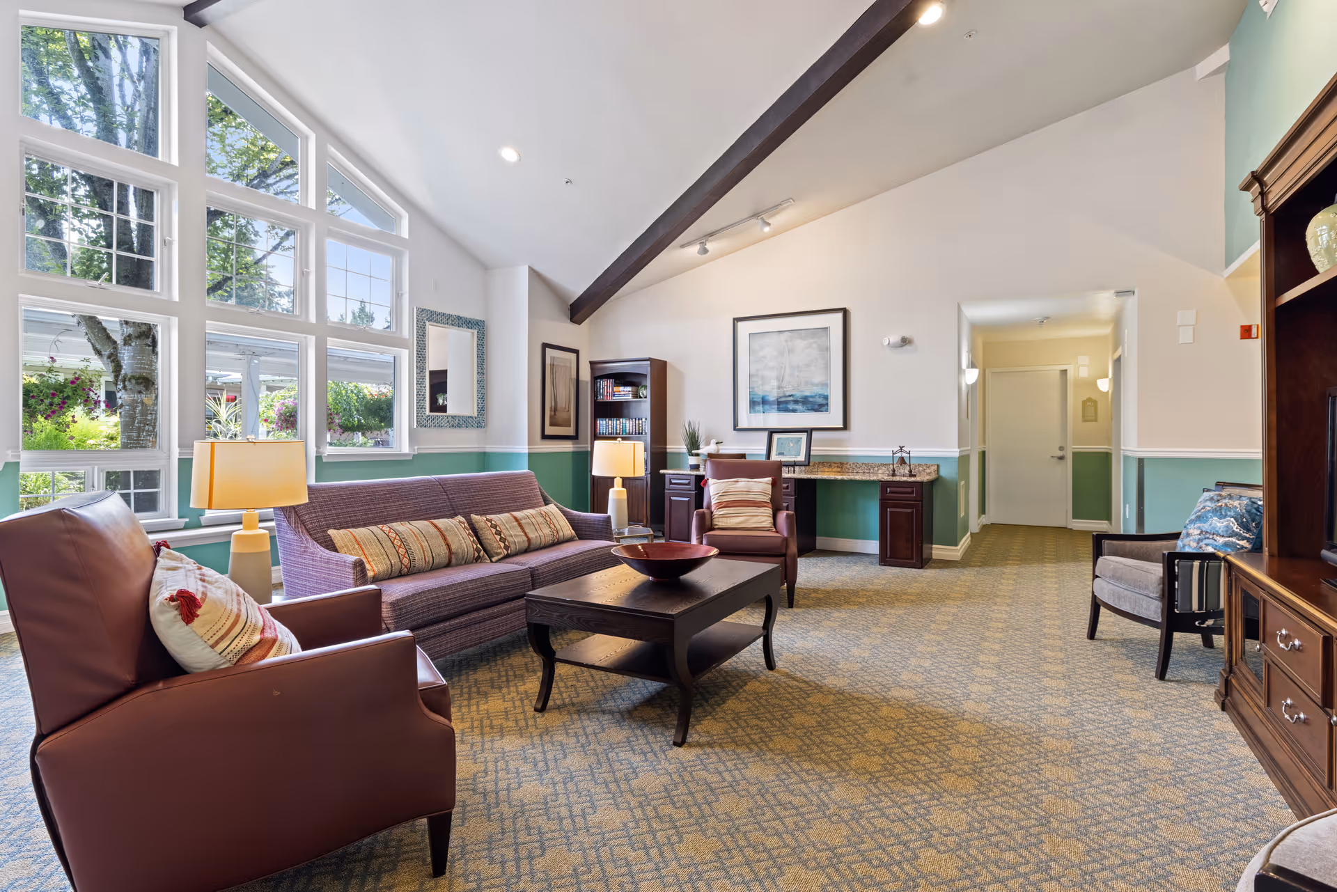 A bright and spacious living room with large windows letting in natural light. The room features a purple sofa with multiple cushions, two leather armchairs, a wooden coffee table with a decorative bowl, and a wooden entertainment unit. The walls are painted white with a green lower half, and there are framed artworks and a mirror on the walls. The carpet has a patterned design, and there are table lamps providing additional lighting.