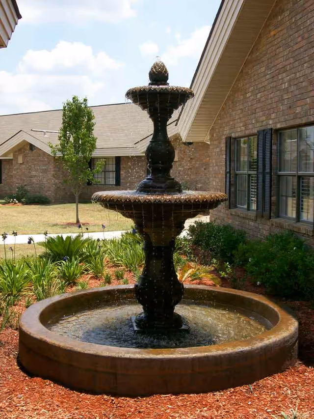 Outdoor view of a tiered water fountain surrounded by mulch and plants, with a brick building and windows in the background under a partly cloudy sky.