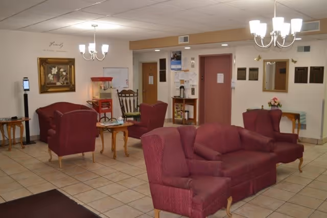 A common room with maroon upholstered sofas and armchairs arranged around small wooden tables, chandeliers overhead, tiled floor, and an elevator/entry area in the background.