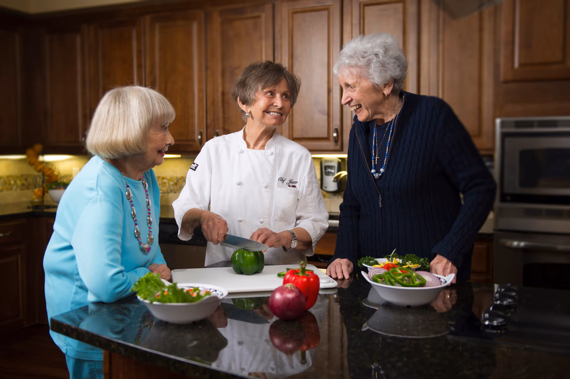 Three older women in a kitchen smiling as one woman in a chef's coat chops a green bell pepper at a countertop with bowls of salad and other vegetables.