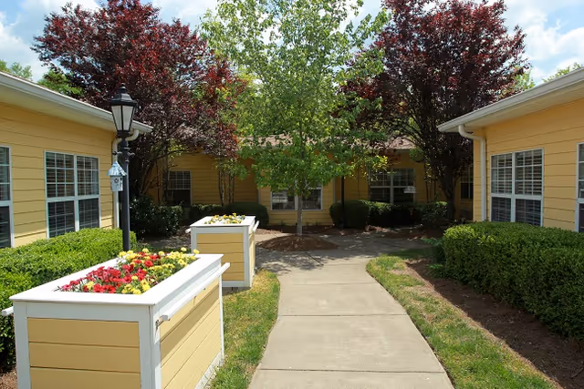 Outdoor courtyard area at Belvedere Commons of Franklin with a concrete walkway flanked by yellow buildings, green bushes, flower planters, and trees under a partly cloudy sky.