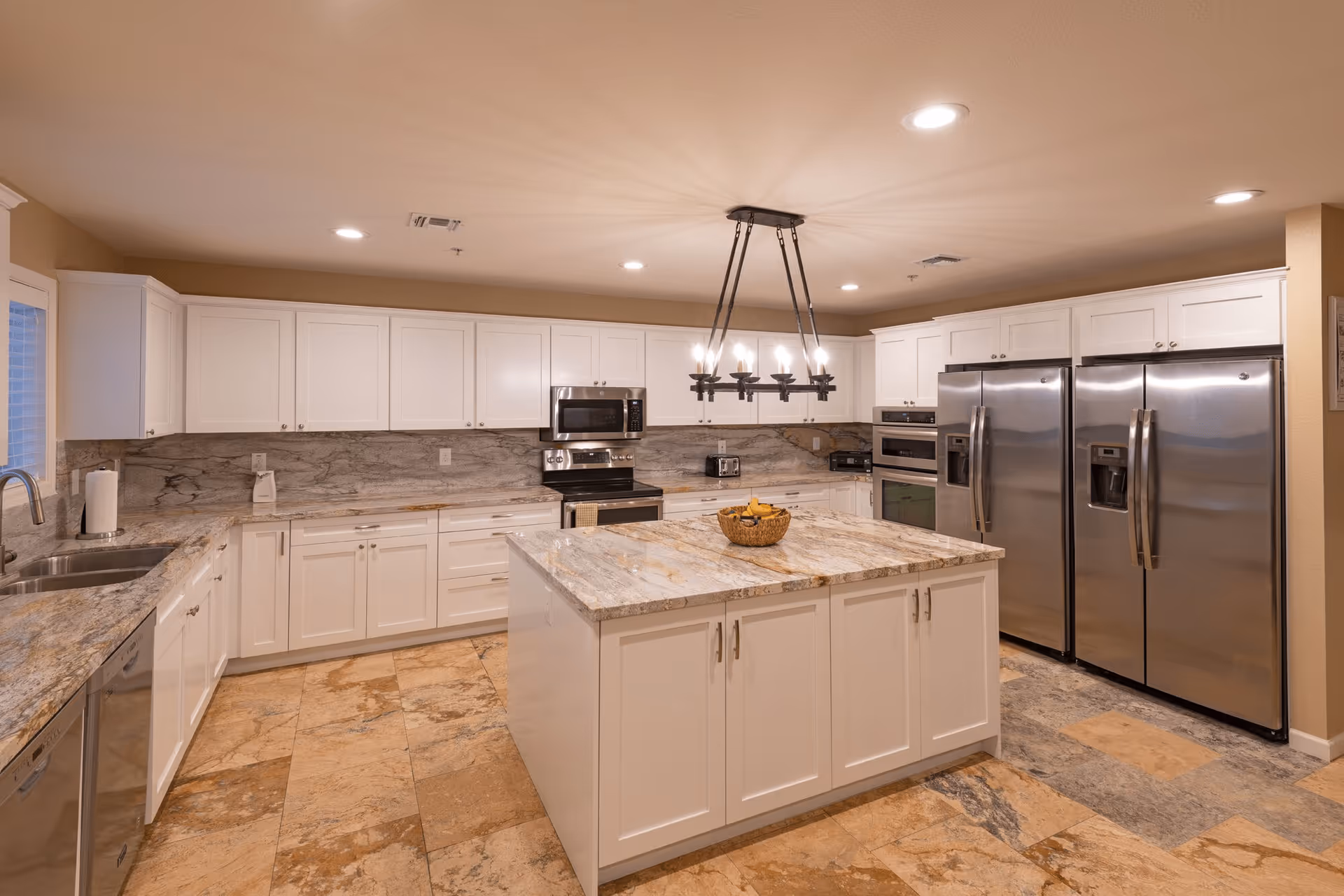 Spacious modern kitchen with white cabinets, granite countertops, a large central island with a basket of bananas, stainless steel appliances including two refrigerators, an oven, microwave, and dishwasher, and a decorative chandelier hanging above the island.