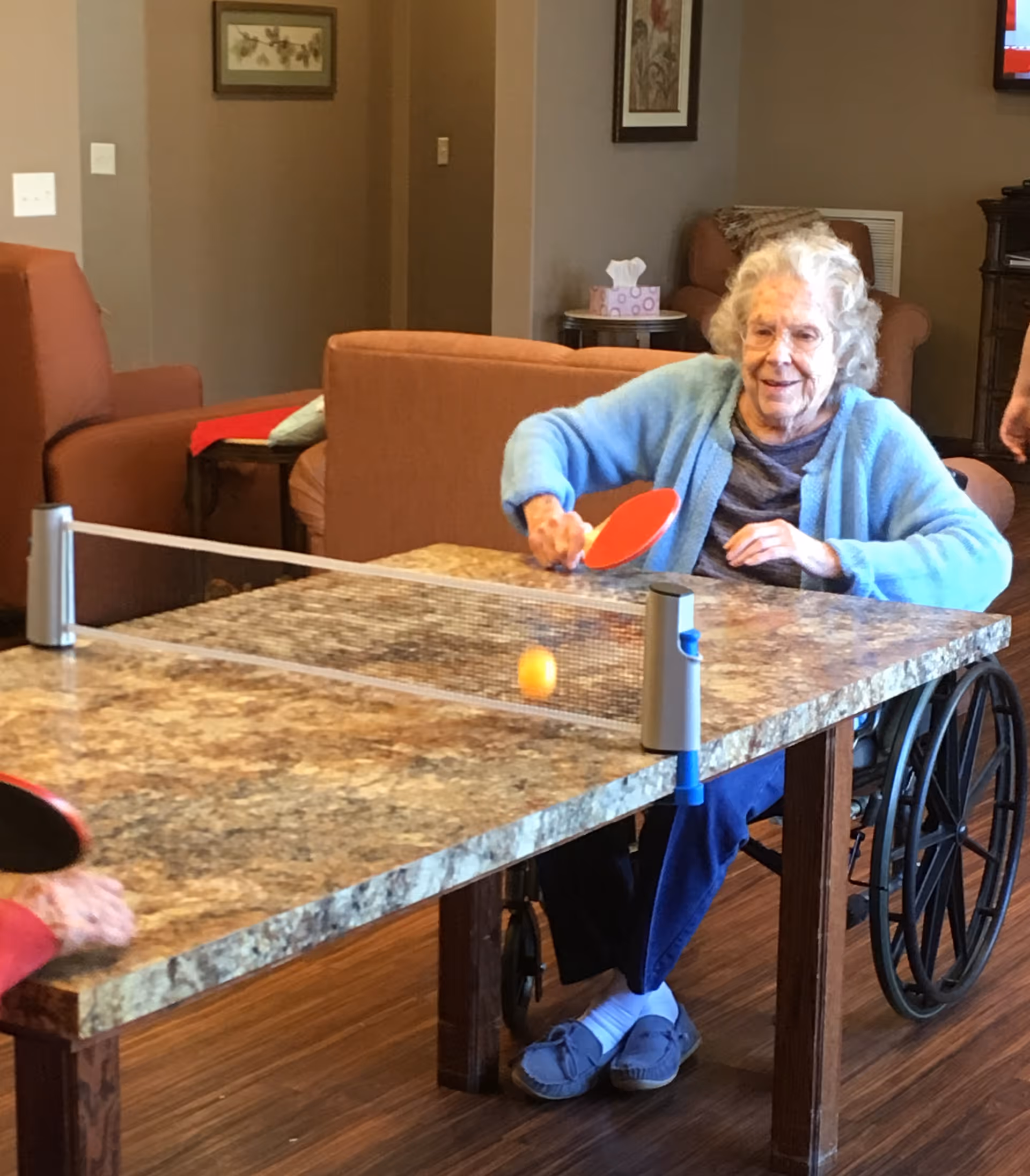 An elderly woman in a wheelchair playing table tennis indoors, hitting an orange ping pong ball with a red paddle across a table with a net. The room has brown armchairs, framed pictures on the walls, and a tissue box on a side table in the background.