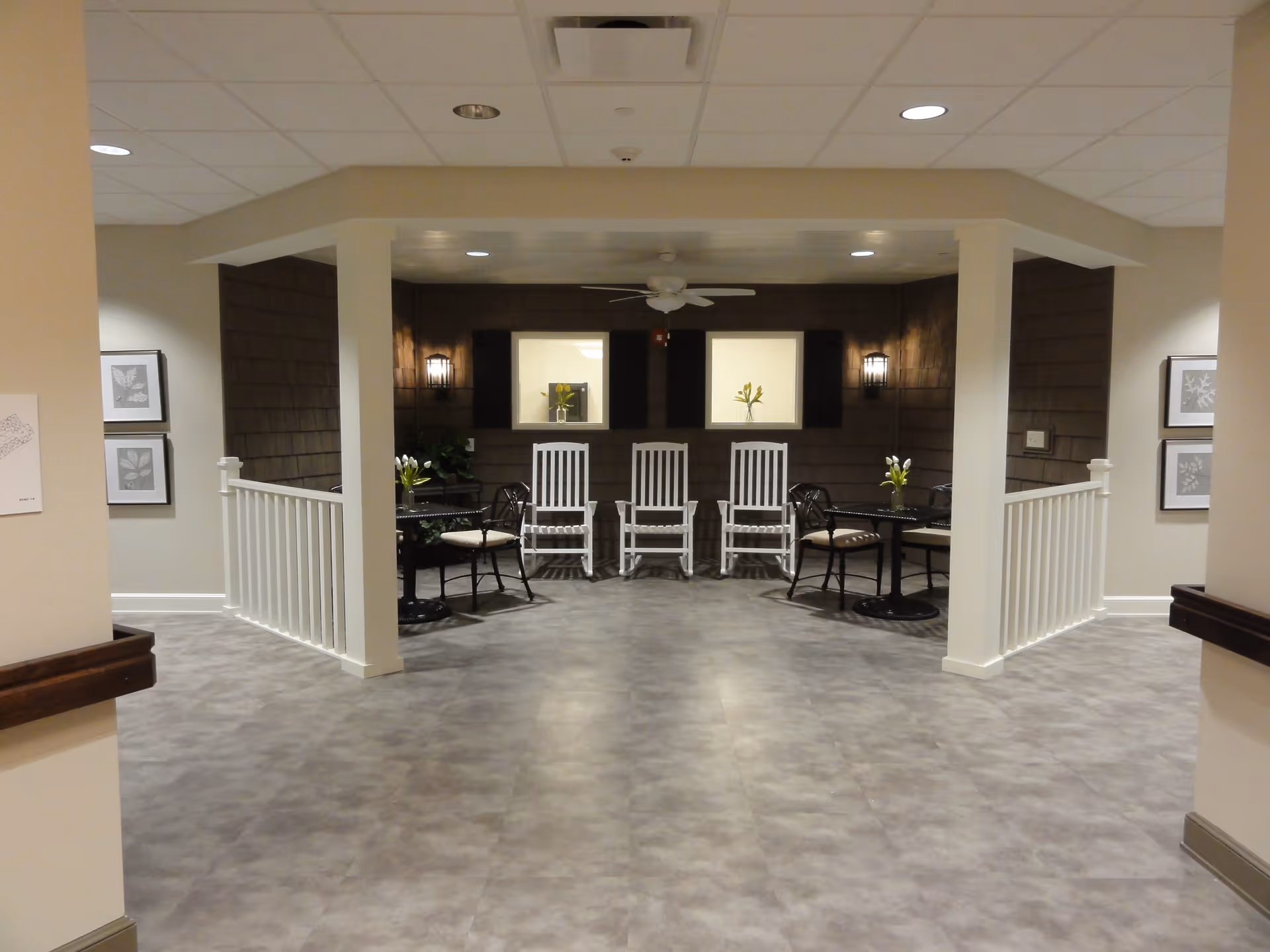 Indoor seating area with three white rocking chairs and two small round tables with black chairs, each table decorated with a small vase of flowers. The space has a ceiling fan, wall sconces, framed artwork on the walls, and a gray floor.