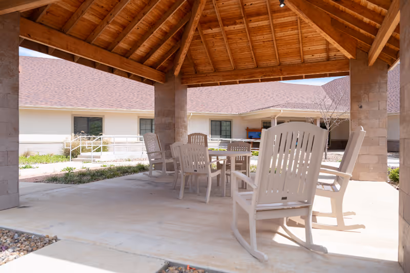 Covered outdoor patio with a wooden vaulted roof, white rocking chairs and a table in a nursing facility courtyard.