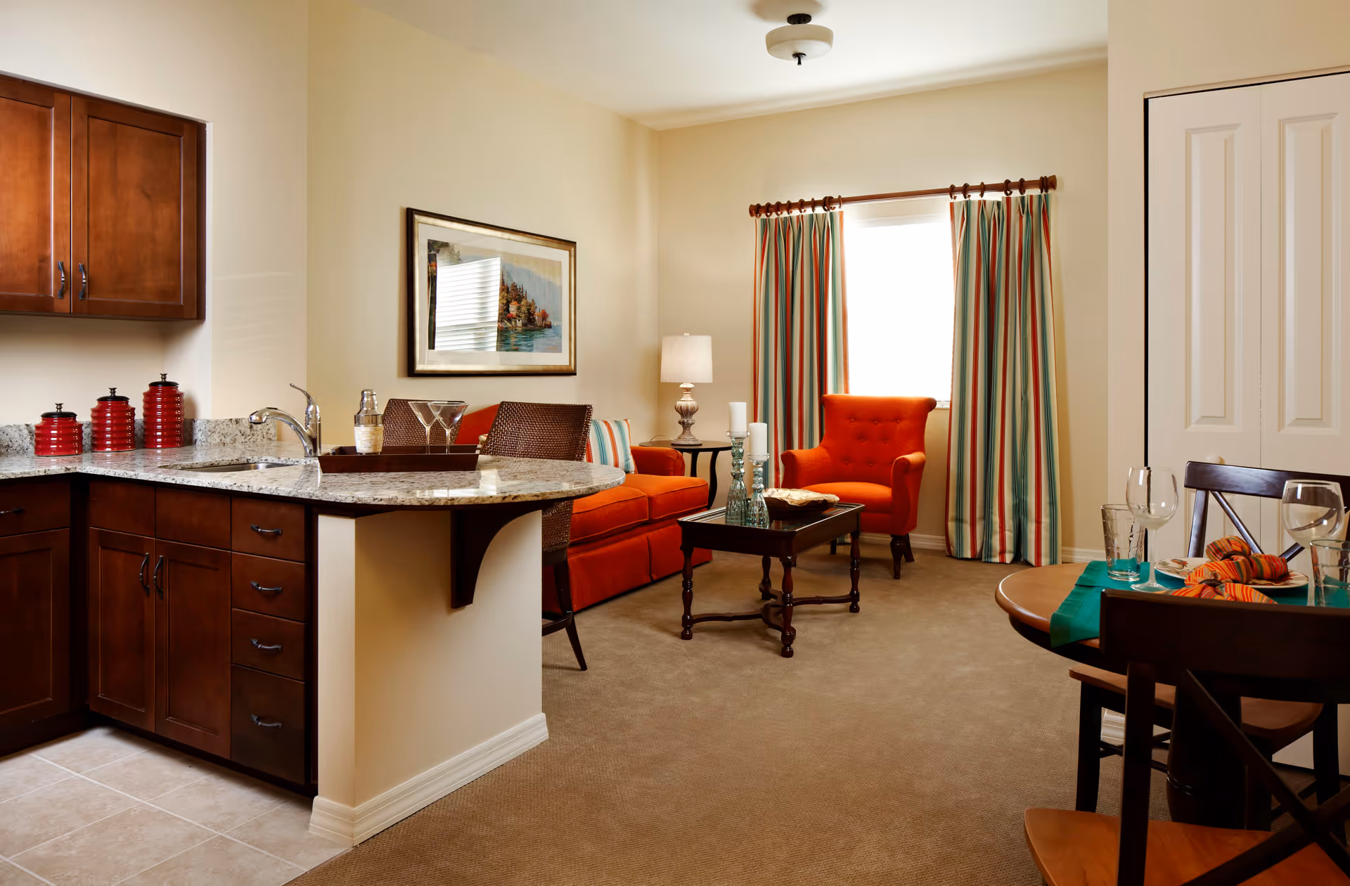 A cozy living area with an orange sofa and matching armchair, a wooden coffee table with candles, and a window with striped curtains. Adjacent to the living area is a kitchen with dark wooden cabinets, granite countertops, and a small dining table set with glasses and napkins.
