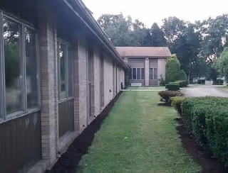 Exterior view of a single-story brick building with multiple windows on the left side and a two-story building in the background. There is a well-maintained lawn with trimmed bushes along the side of the building and trees in the distance.