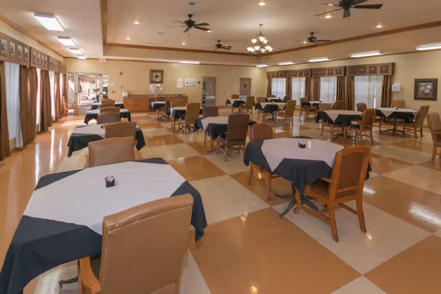 Spacious dining room with multiple tables draped in tablecloths and chairs arranged on a checkered tile floor under ceiling fans and chandeliers.