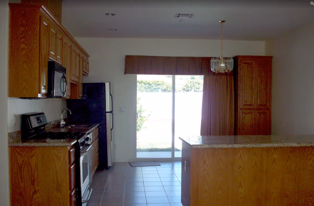 Interior view of a kitchen with wooden cabinets, a black refrigerator, a stove with an oven, a microwave, a sink, and a granite countertop island. There is a sliding glass door with brown curtains leading outside.