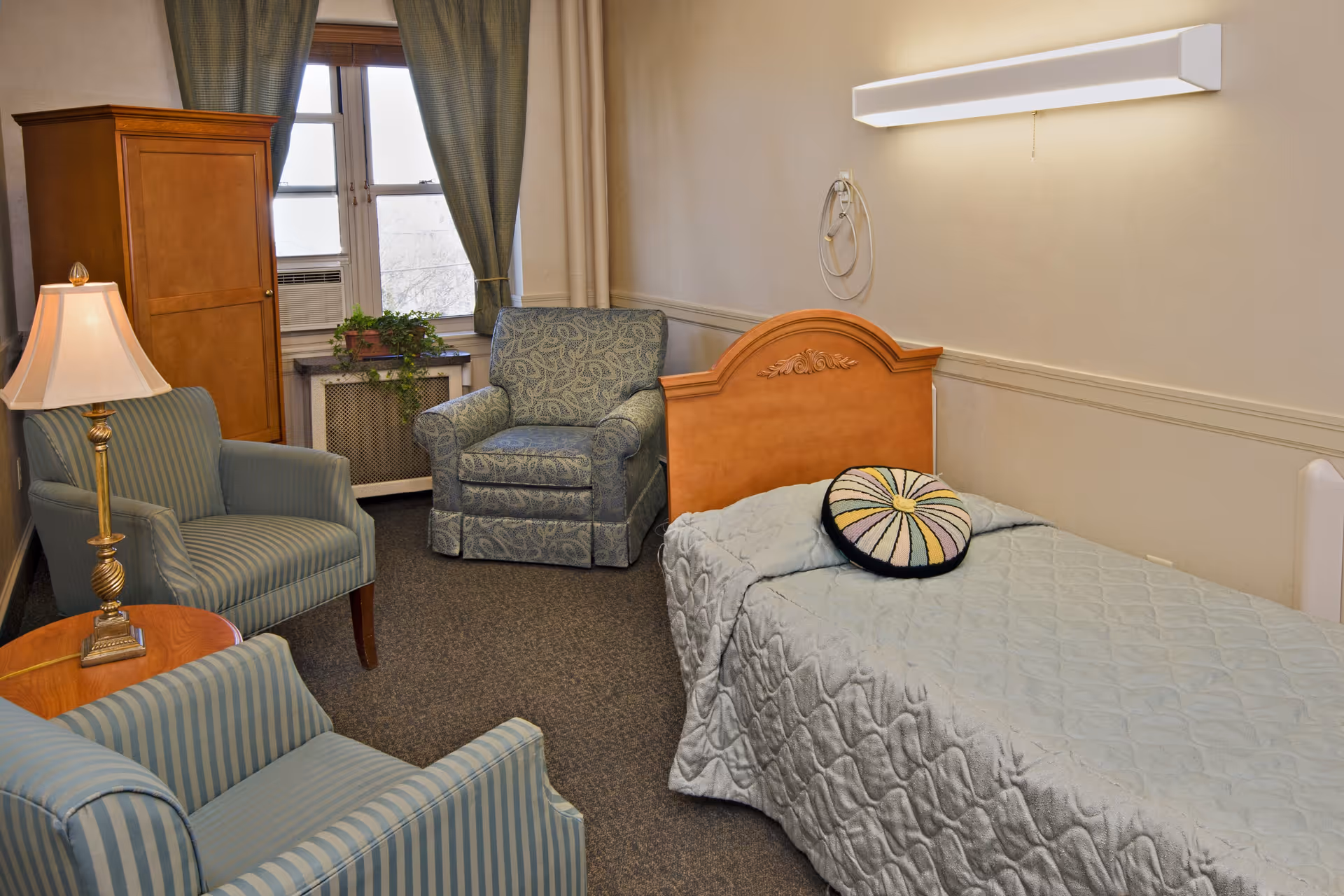 A cozy room with a single bed covered in a light blue quilt and a round decorative pillow. There are two striped armchairs and one patterned armchair arranged around a small wooden table with a lamp. A wooden cabinet stands near a window with green curtains and a potted plant on the windowsill.