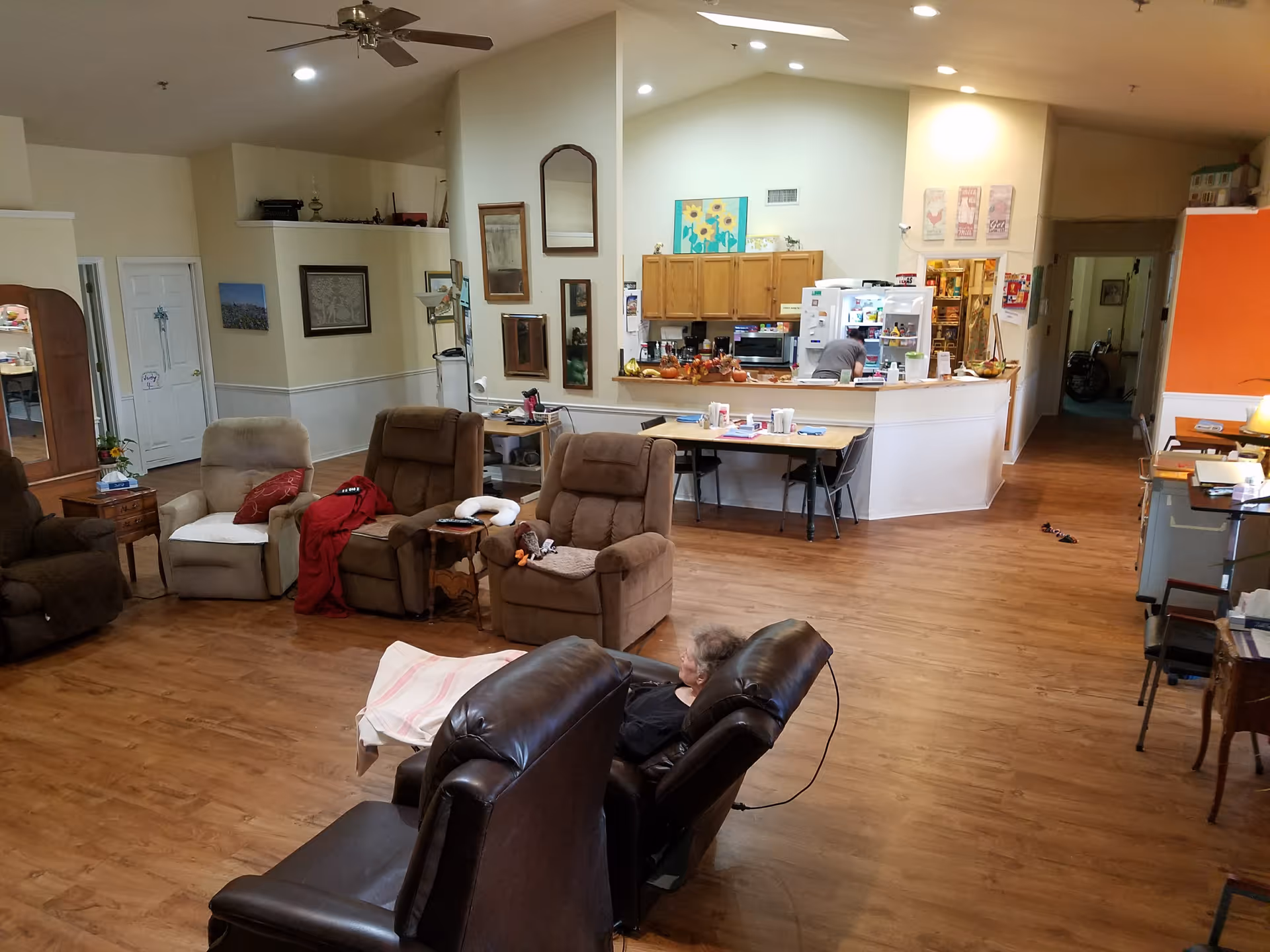 Open assisted-living common room with several recliner chairs, a person seated in a recliner, and a kitchenette/dining counter in the background.