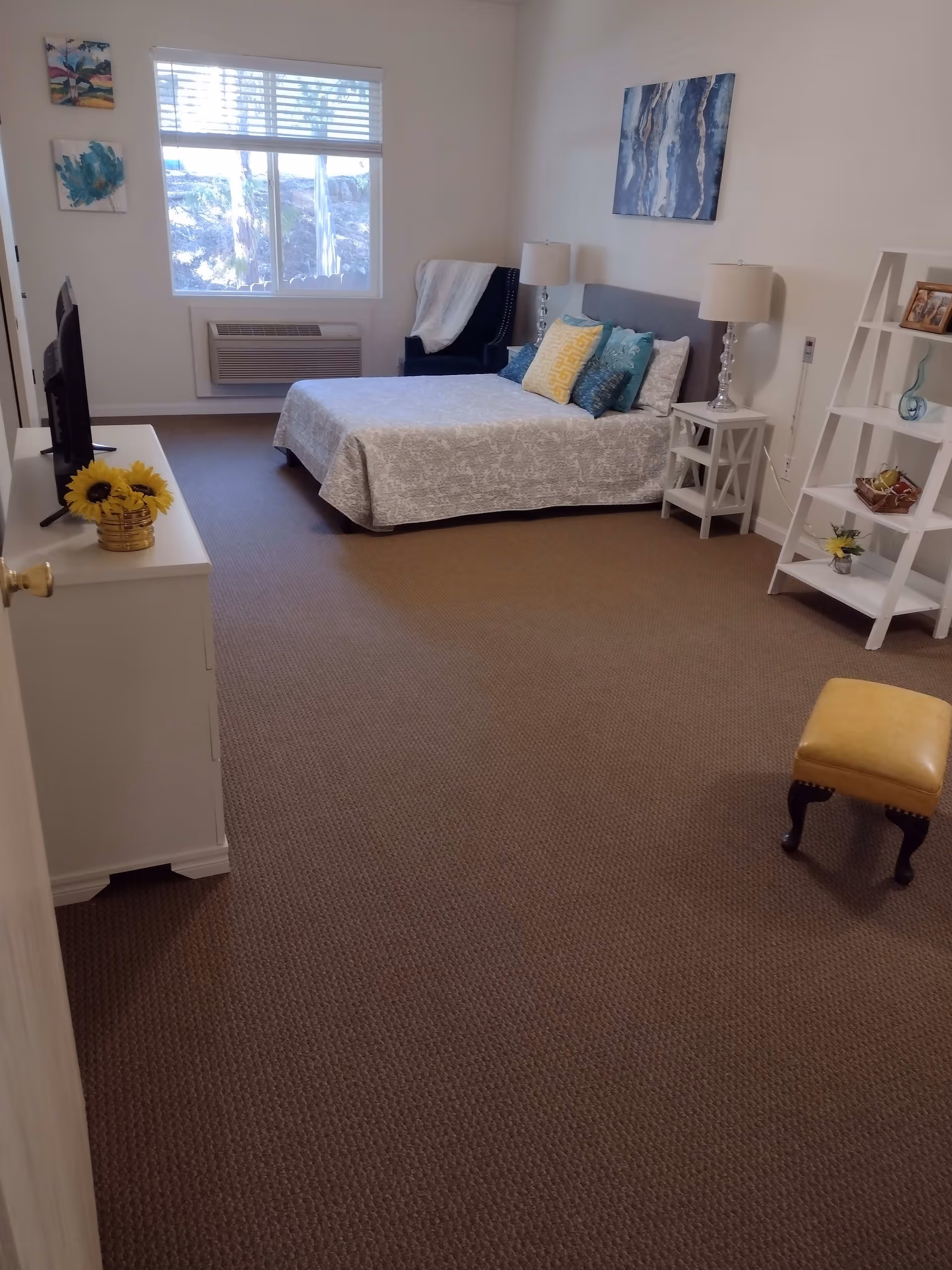 A bedroom with a bed covered in a light gray patterned bedspread and multiple decorative pillows. There are two bedside tables with lamps, a dark armchair with a white throw blanket near a window, a white shelving unit with decorative items, a small yellow ottoman, and a white dresser with a TV and a vase of sunflowers. The room has beige carpet and white walls with two paintings.