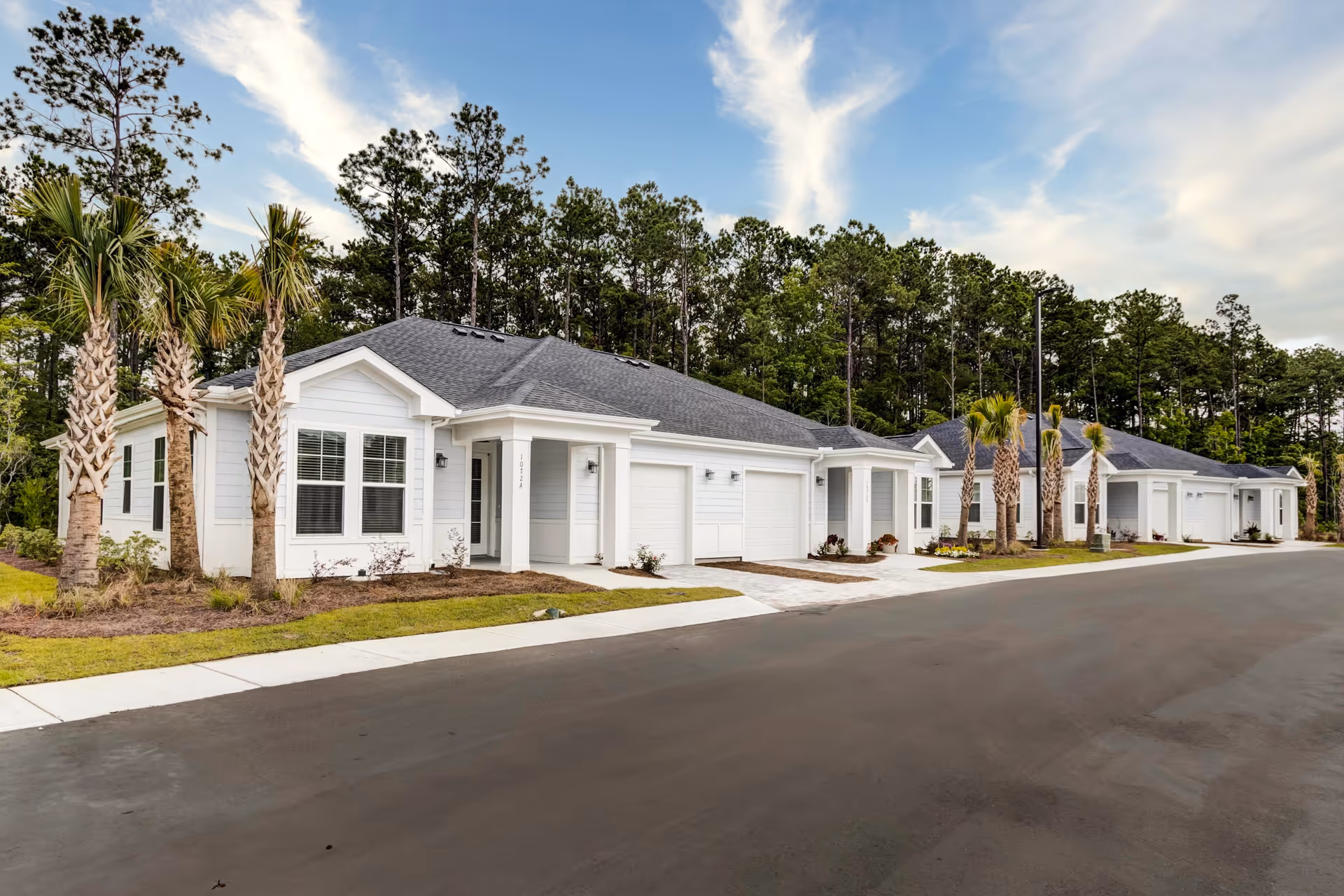 Row of single-story white residential buildings with garages, palm trees, and a paved road in front, set against a backdrop of tall pine trees under a partly cloudy sky.