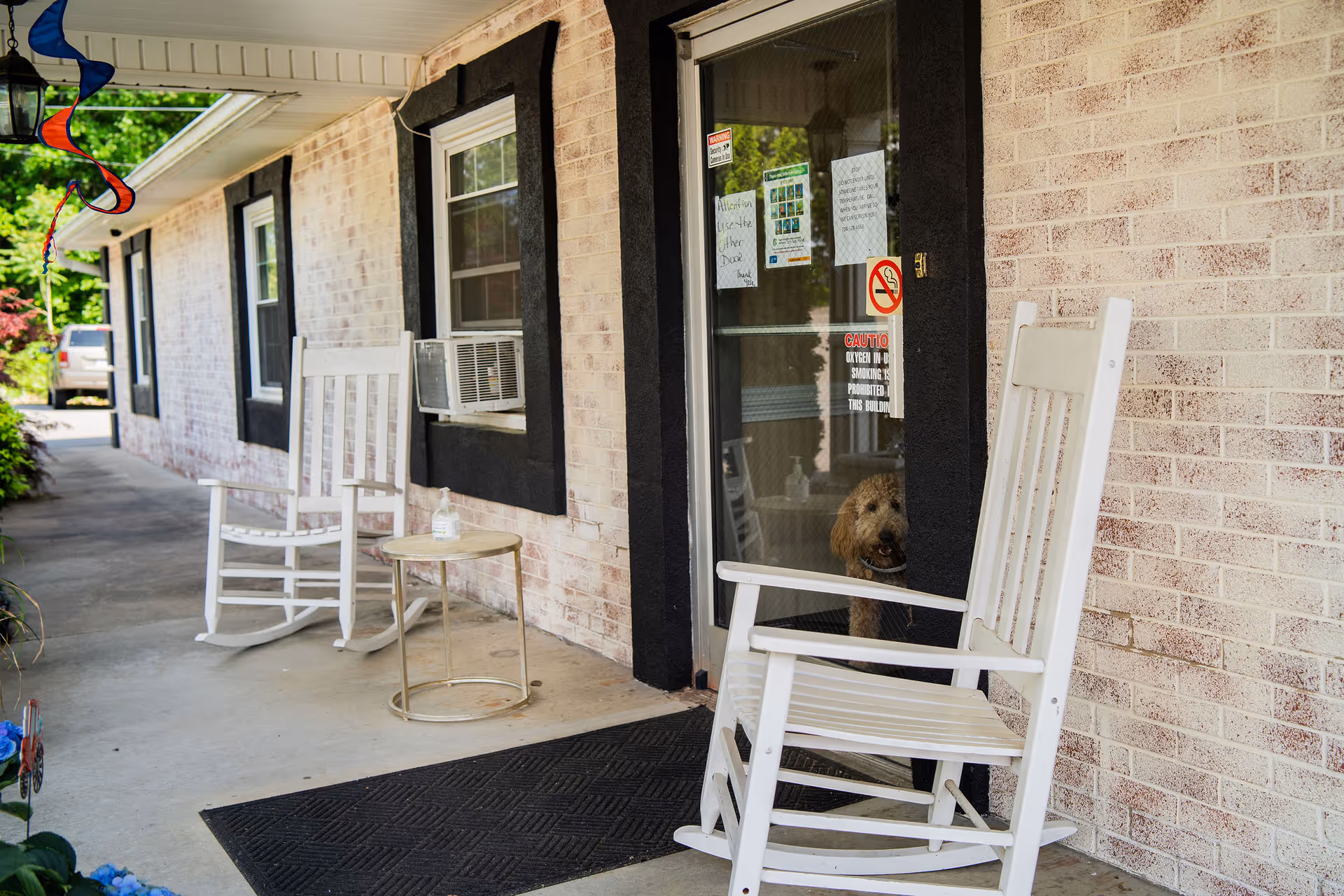 Front porch of a brick memory care facility with white rocking chairs, a small table, and a dog peeking through a glass door.