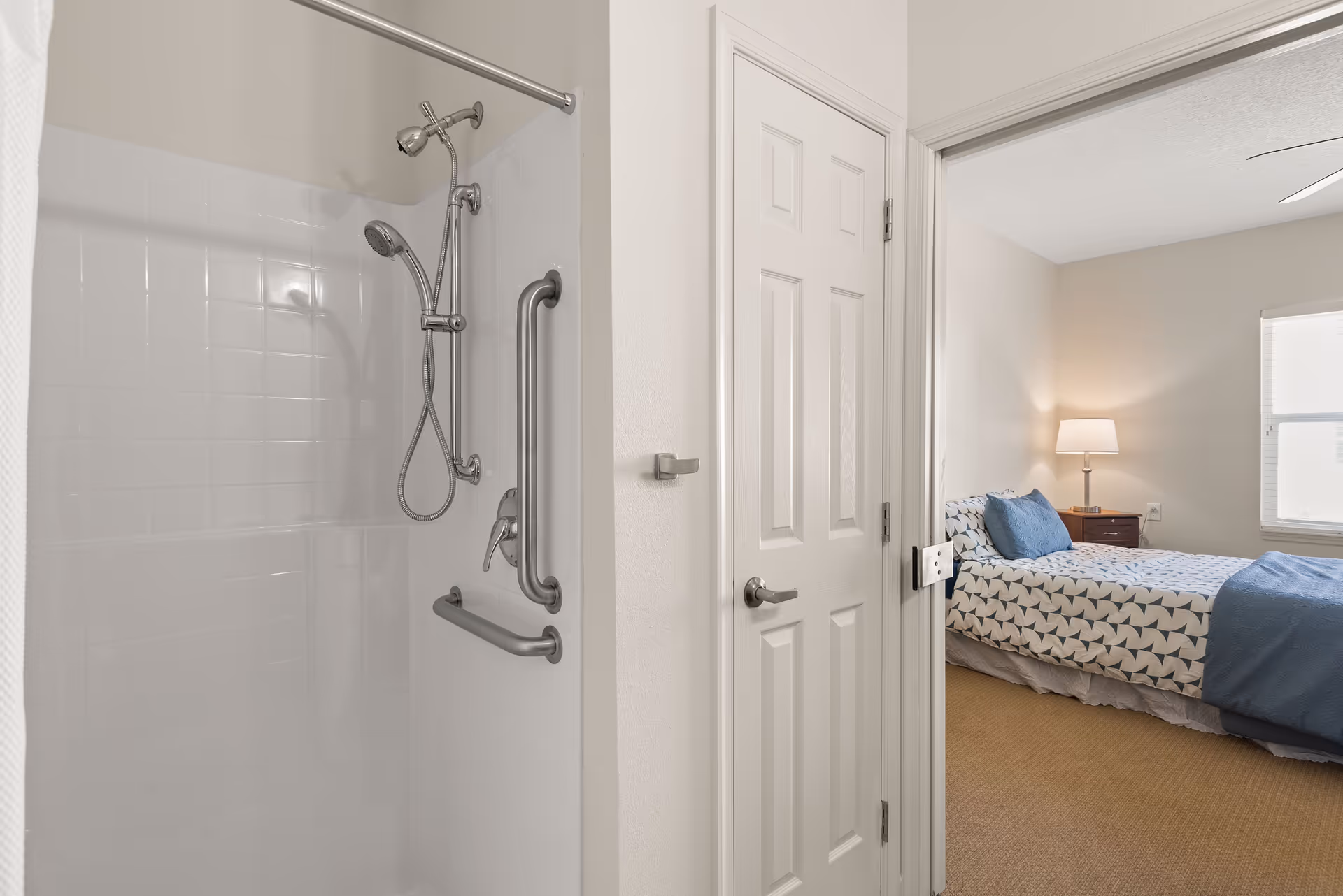 View of a bathroom shower with white tiled walls and metal grab bars, adjacent to a bedroom with a bed covered in patterned bedding, a nightstand with a lamp, and a window with blinds.