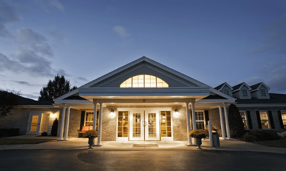 Front exterior view of a senior living facility named Tennyson Court at dusk, showing a well-lit entrance with double glass doors, white columns, and a triangular pediment with a large window above. There are potted plants on either side of the entrance and windows with black shutters on the building.